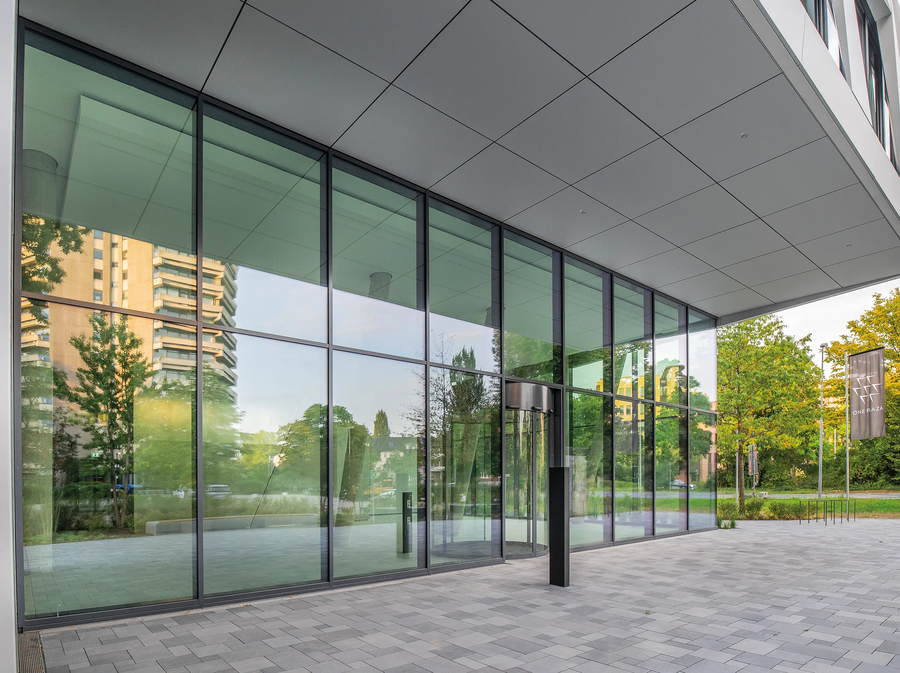 Ingangsgebied van ONE PLAZA met een royale glazen gevel, moderne architectuur en uitzicht op de omliggende gebouwen en groene ruimtes.